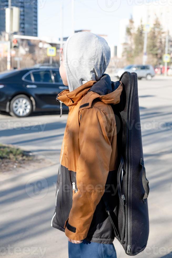 un' adolescenziale ragazzo nel un' giacca e cappello passeggiate solo con un' chitarra nel un' Astuccio. bambino musicista perso nel il città foto