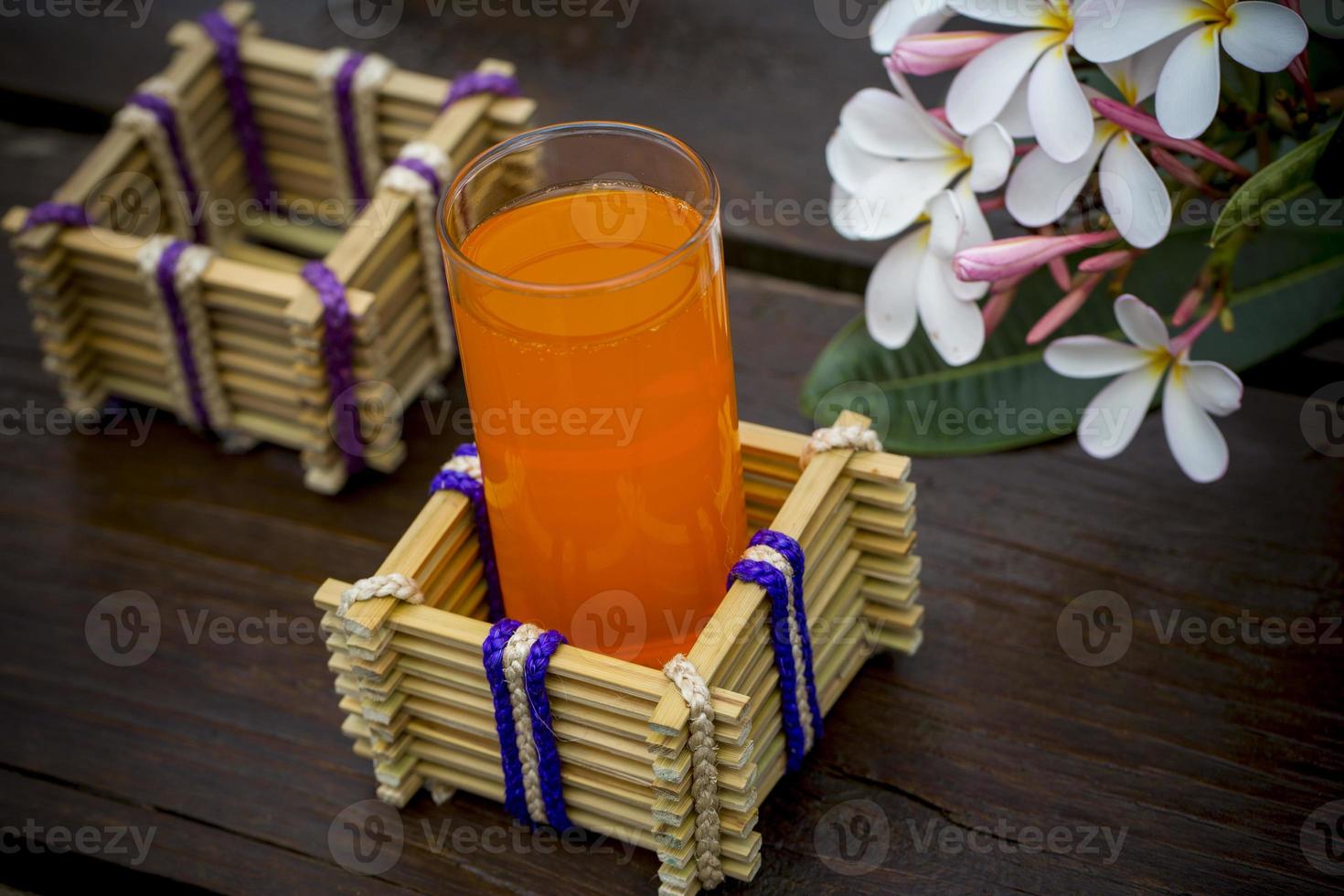 un' bicchiere di arancia succo con acqua bicchiere titolare fatto di bambù bastoni e fibra. decorativo di legno tavolo con bellissimo plumeria fiori. bambù bicchiere titolare. foto