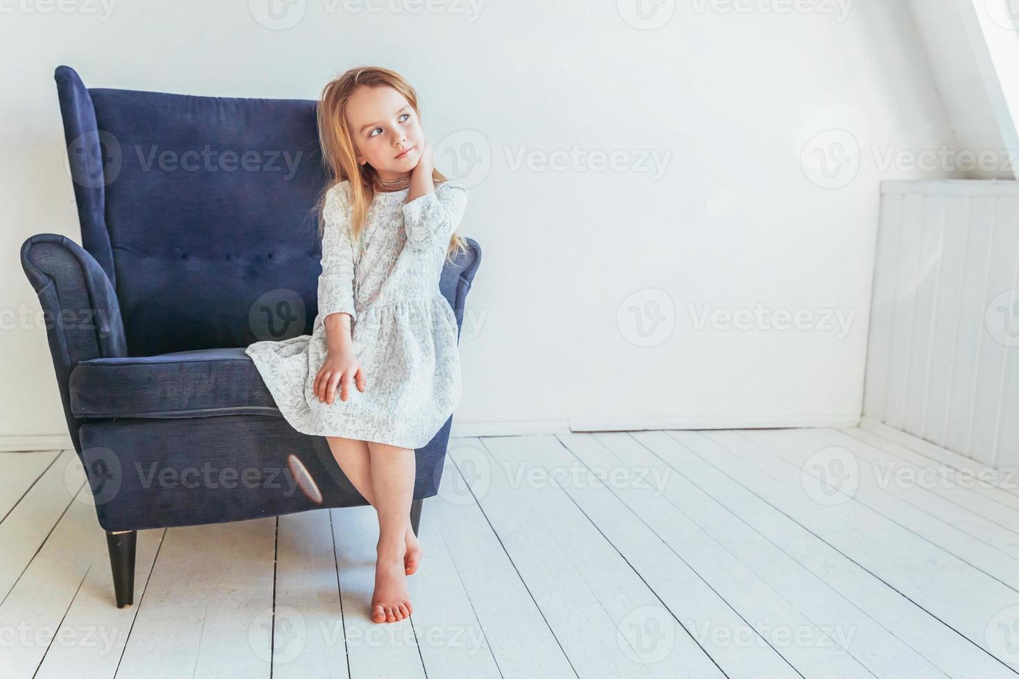 dolce poco ragazza nel bianca vestito seduta su moderno accogliente blu sedia rilassante nel bianca luminosa vivente camera a casa al chiuso. infanzia scolari gioventù rilassare concetto. foto