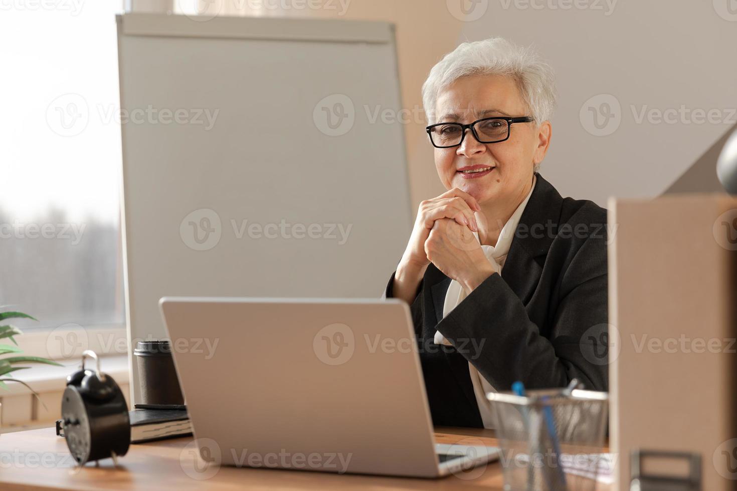 ritratto di fiducioso elegante europeo mezzo anziano anziano donna a posto di lavoro. elegante più vecchio maturo 60s grigio dai capelli signora donna d'affari seduta nel moderno ufficio. capo capo insegnante professionale lavoratore. foto