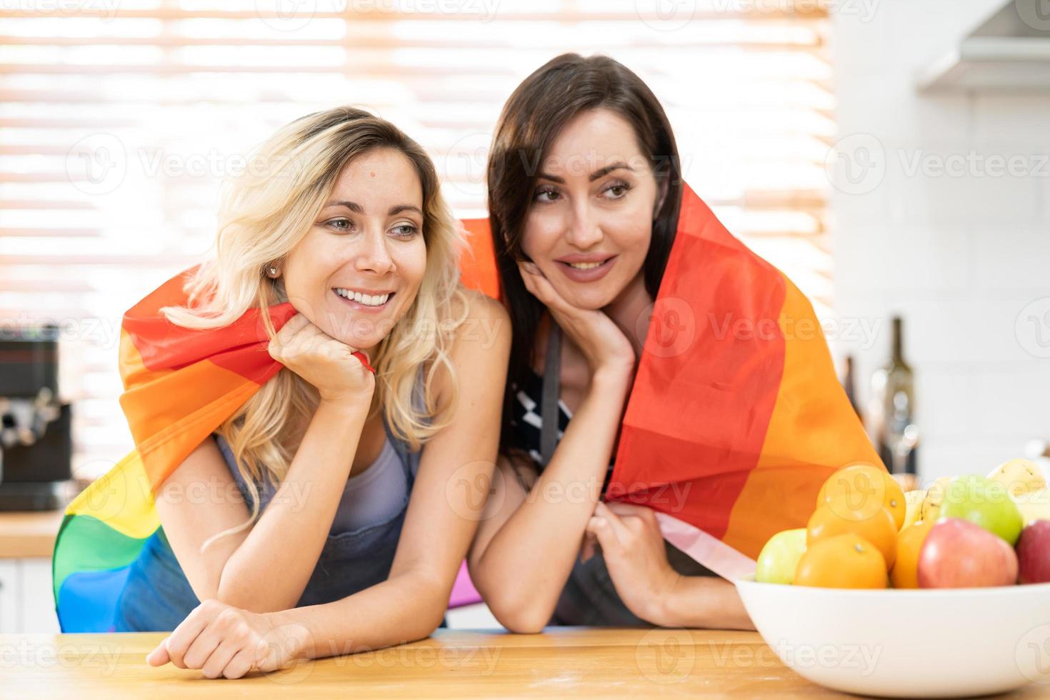 contento caucasico lgbt coppia, lesbica Tenere e agitando arcobaleno lgbt orgoglio bandiera insieme nel il cucina a casa. diversità di lgbt relazioni. allegro omosessuale coppia. foto