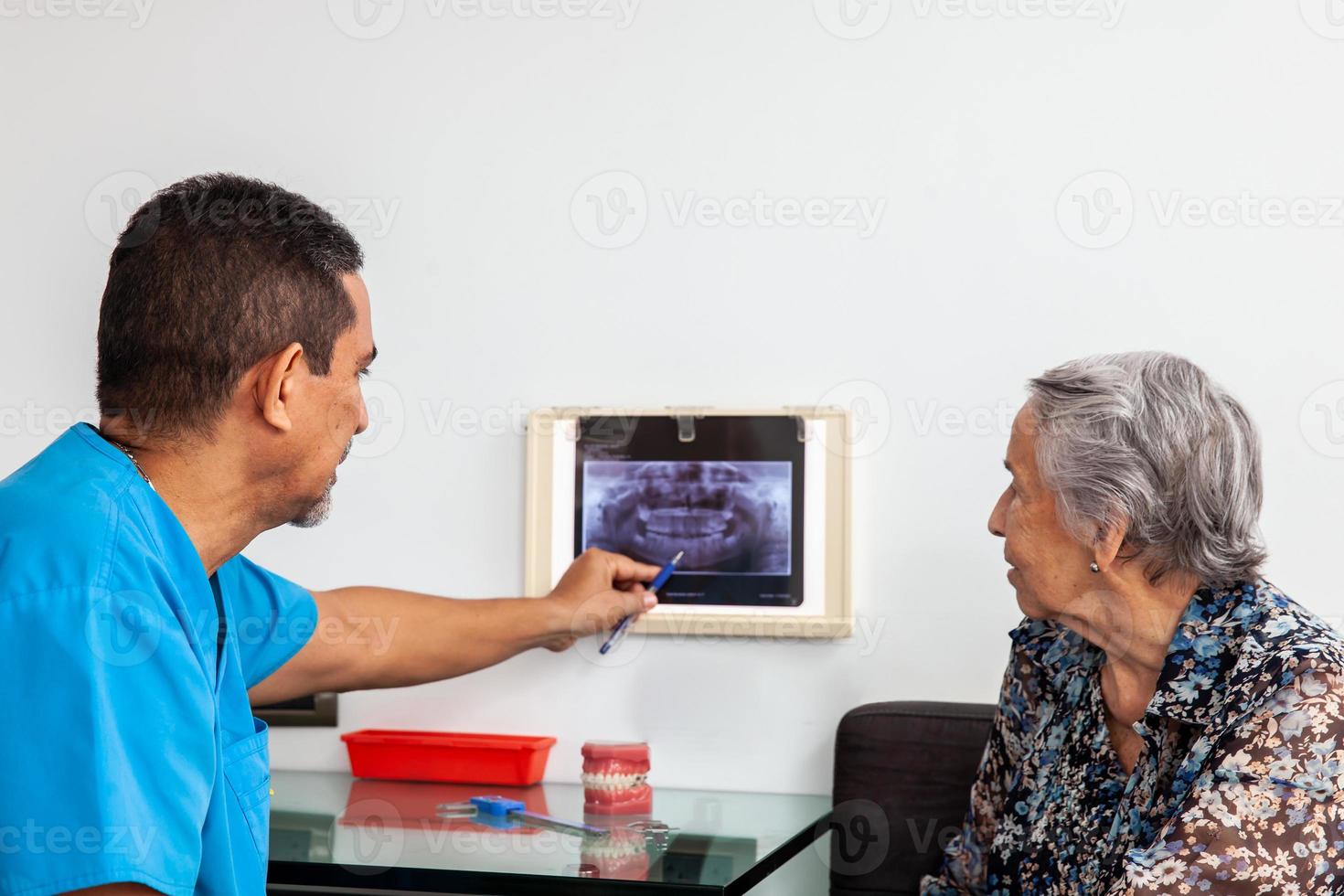 maschio mezzo -invecchiato dentista a il suo ufficio utilizzando un X raggio Immagine per spiegare trattamento per il suo anziano donna paziente foto