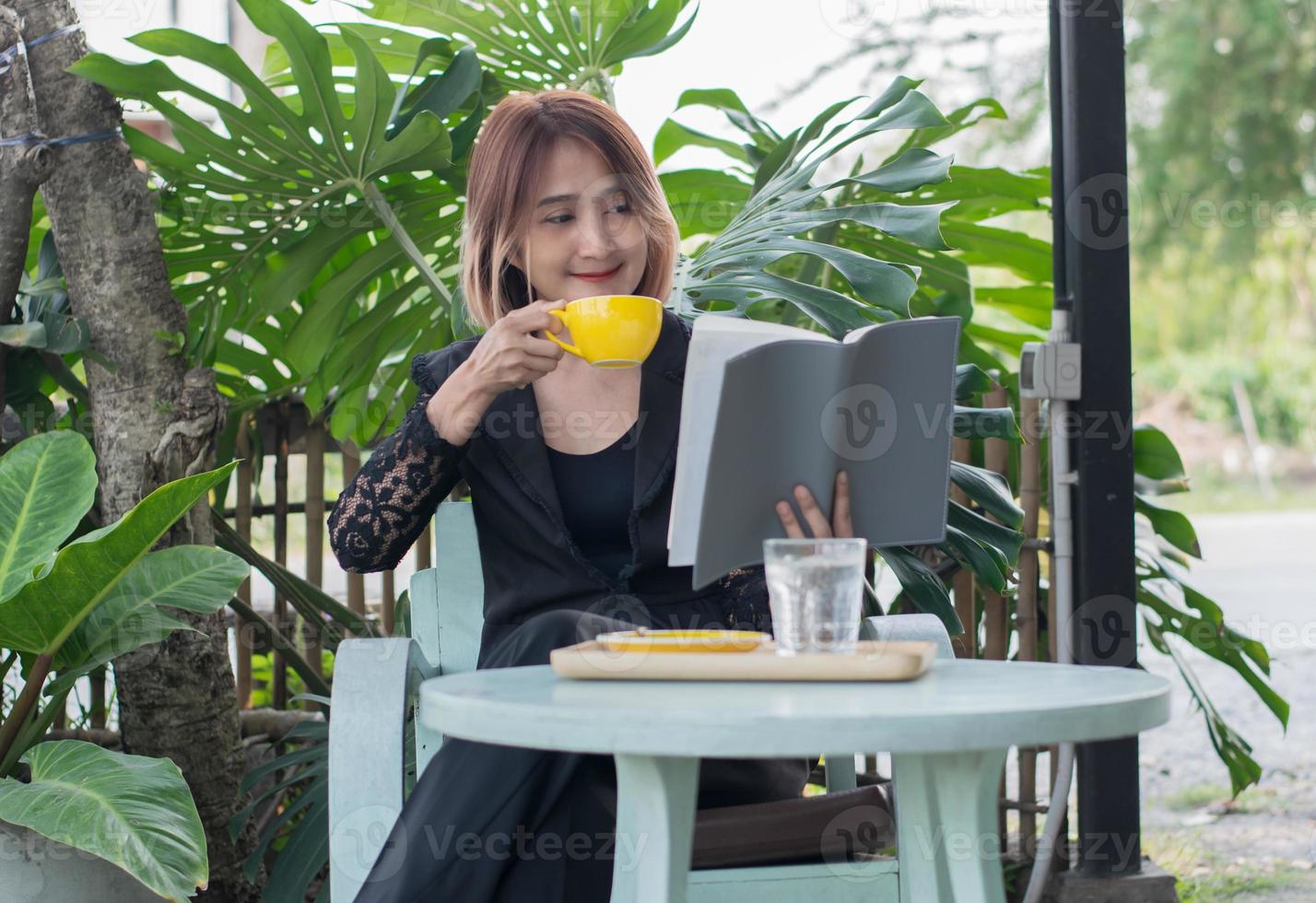 libero professionista donna seduta su un' di legno sedia rilassante nel il giardini ansimare a casa Tenere un' caffè tazza e lettura un' libro per apprendimento con contento sensazione e stile di vita concetti. foto