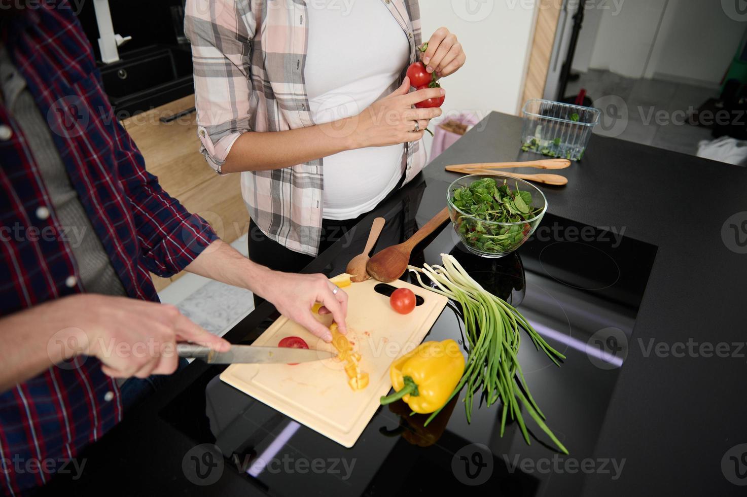 avvicinamento di mani chopping verdure su un' taglio tavola e ritagliata Visualizza per un' incinta donna Tenere rametto con fresco pomodori mentre godendo cucinando insieme con sua marito a casa cucina foto
