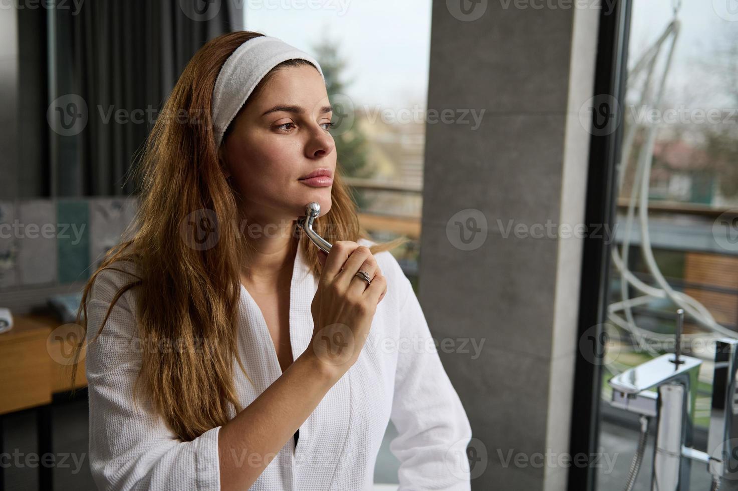 avvicinamento bellissimo sereno incinta europeo donna seduta nel un' bianca accappatoio su il bordo di il bagno e massaggio sua viso con un' rullo massaggiatore, guardare su il finestra prospiciente il giardino foto