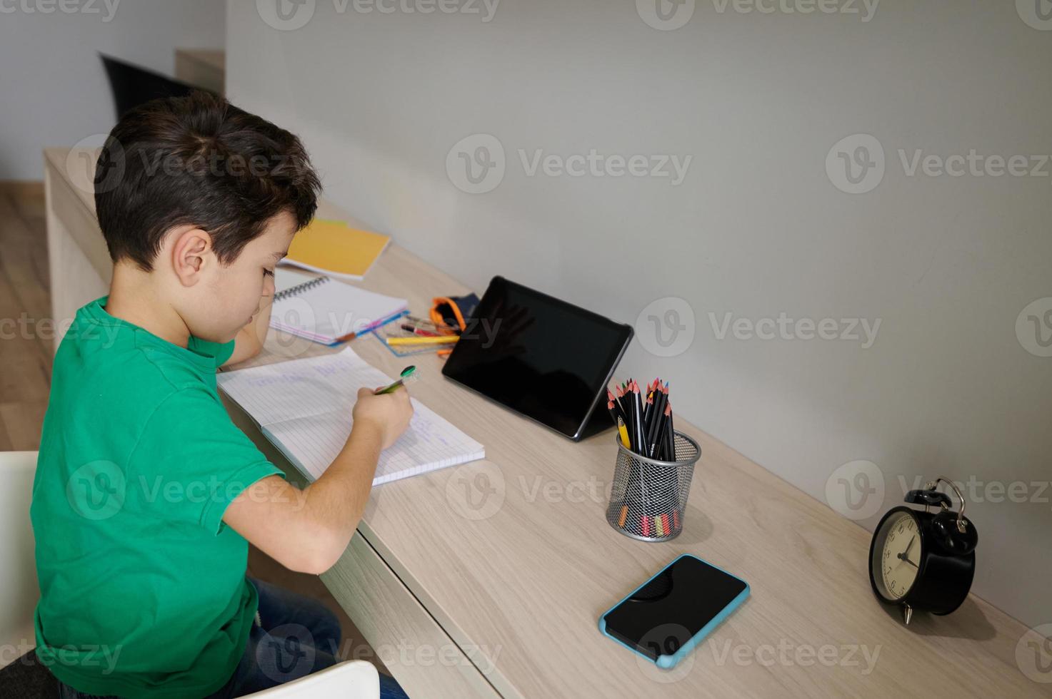 preadolescente scolaro, intelligente bambino studiando a casa, fare il suo compiti a casa, seduta a un' di legno scrivania nel davanti di un' digitale tavoletta. in linea apprendimento, formazione scolastica e conoscenza concetto. foto