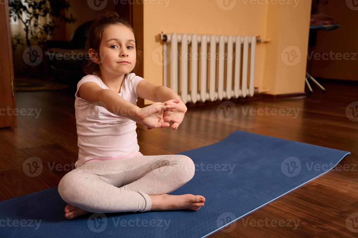 caucasico bello ragazzo ragazza 5 anni vecchio, seduta a piedi nudi su yoga stuoia, allungamento braccia, guardare con fiducia a telecamera foto