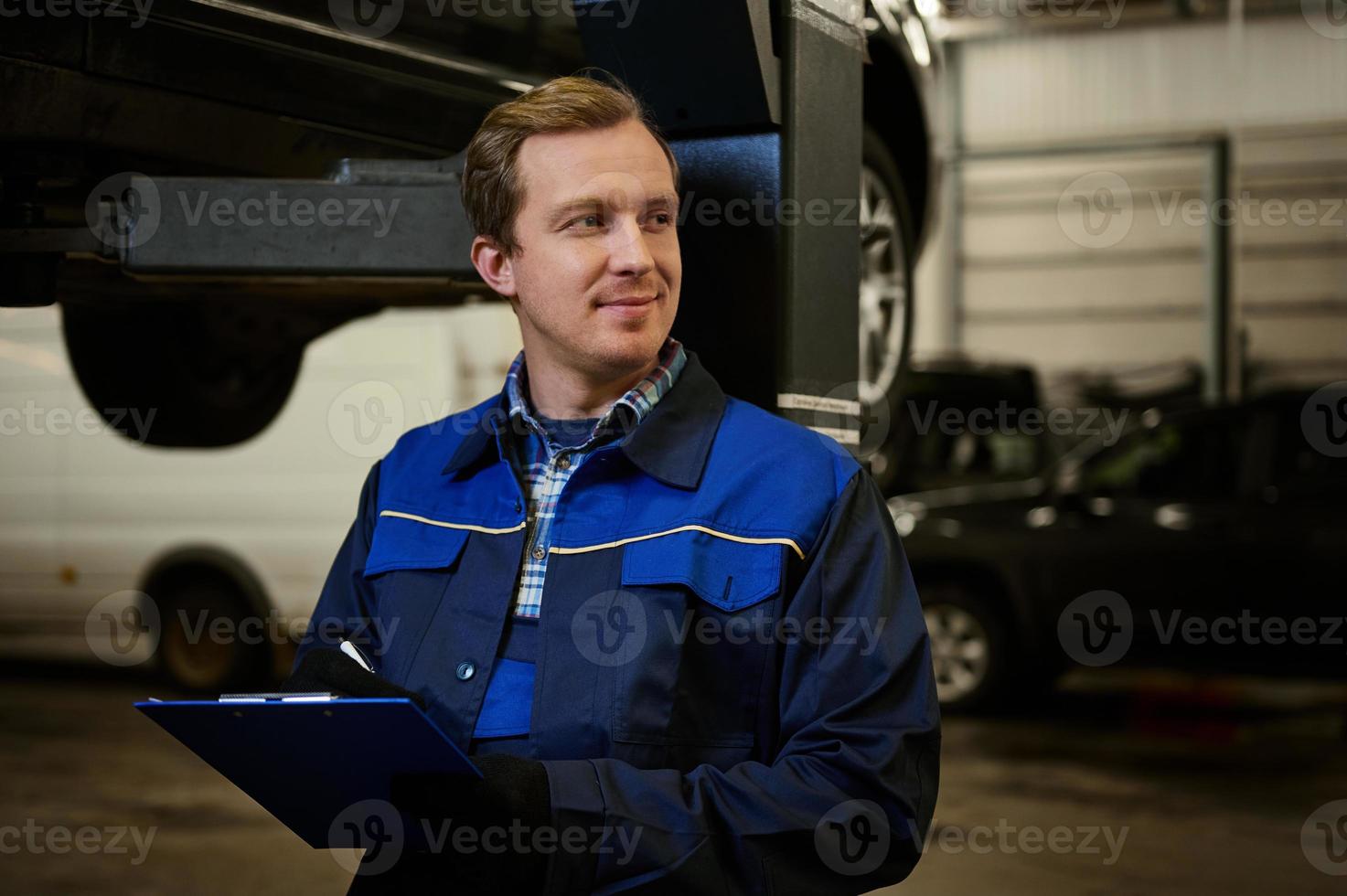 ritratto di un' caucasico auto meccanico, tecnico, auto ingegnere nel uniforme fabbricazione il lista di controllo su appunti per riparazione il auto sollevato su un' montacarichi nel il riparazione negozio durante garanzia auto Manutenzione. foto