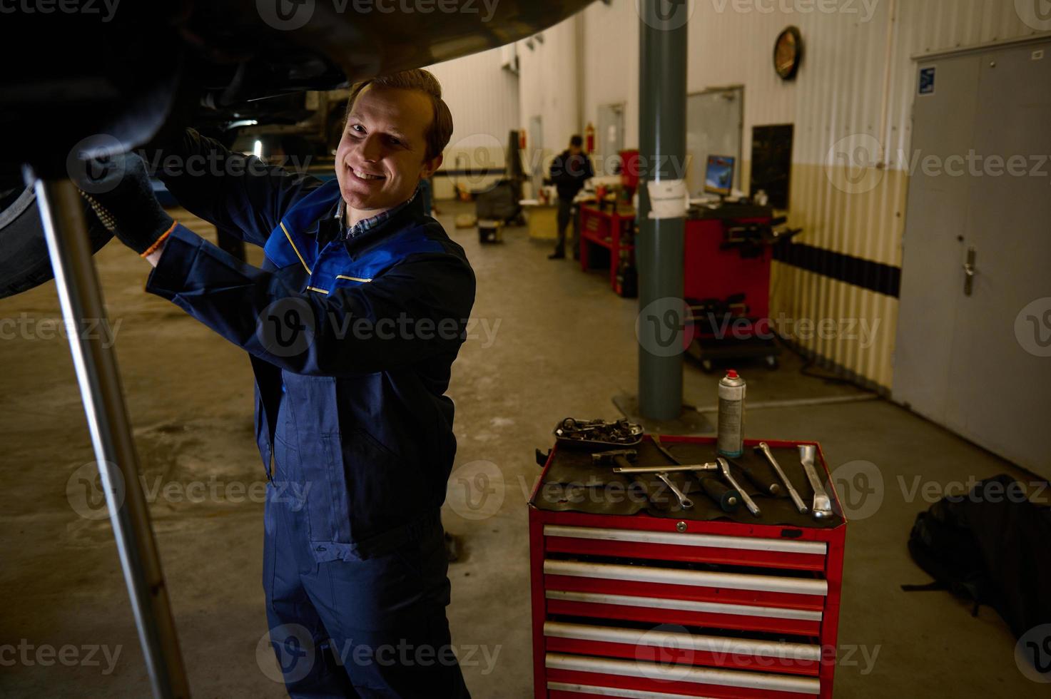 bello caucasico giovane uomo, auto ingegnere, tecnico, auto meccanico riparazione un' sollevato moderno automobile su un' montacarichi nel il riparazione negozio box auto. automobile Manutenzione e auto servizio concetto foto