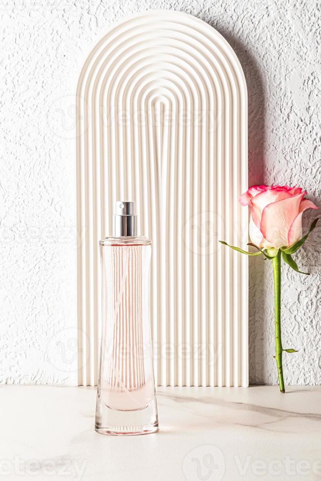 un' elegante alto bottiglia di Da donna profumo contro il sfondo di un' bianca decorativo arco con un' rosa fiore. verticale Visualizza. vuoto bottiglia. foto