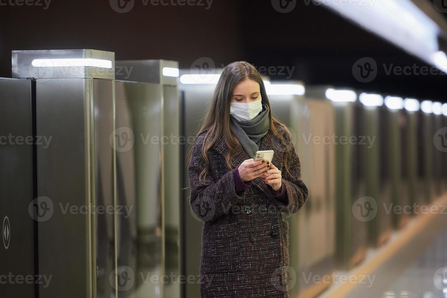 una donna con una maschera medica sta aspettando un treno e tiene in mano uno smartphone foto