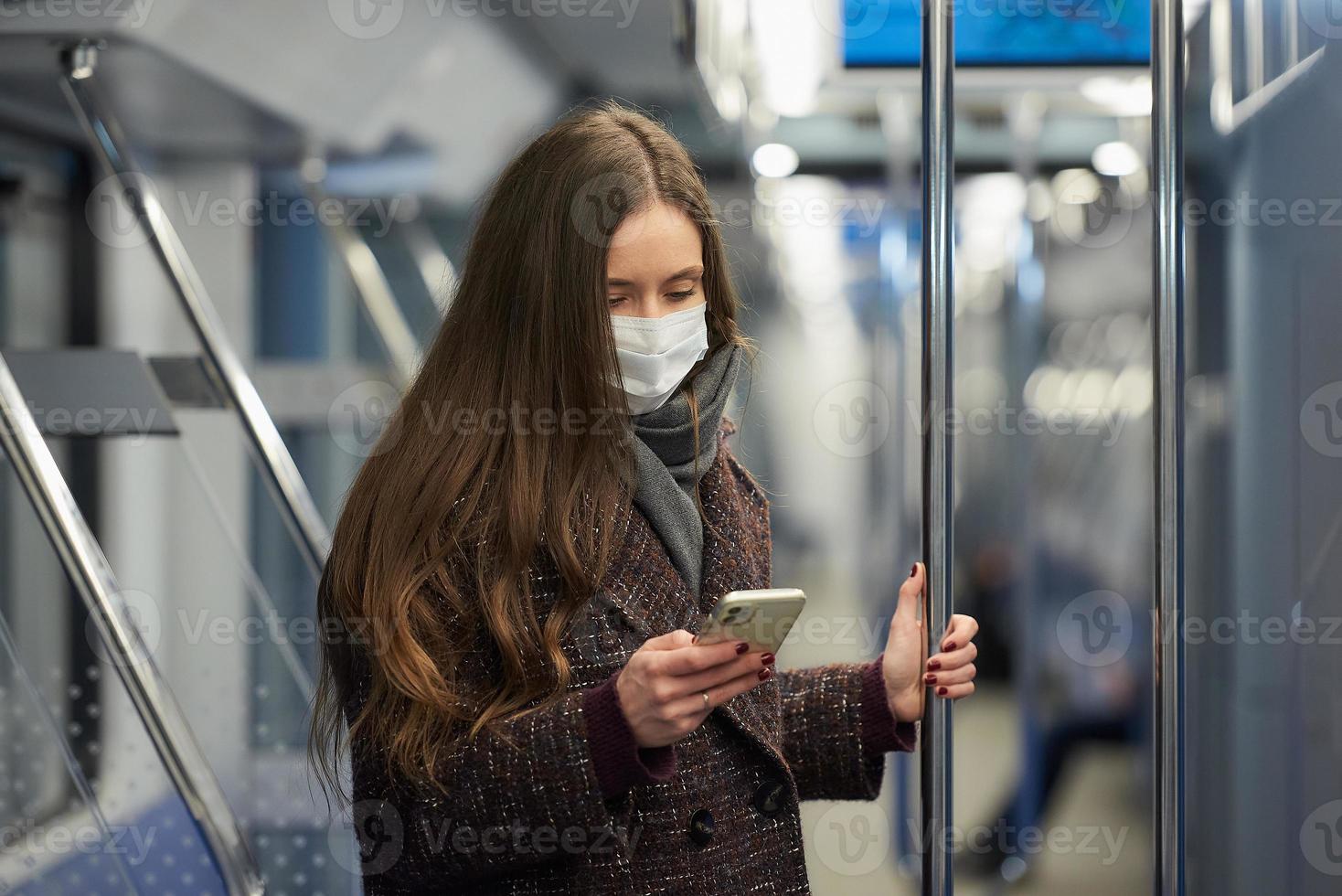 una donna con una maschera è in piedi e utilizza uno smartphone in un moderno vagone della metropolitana foto