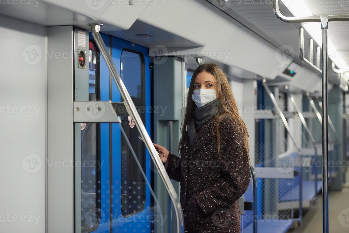 una donna con una maschera è in piedi e utilizza uno smartphone in un moderno vagone della metropolitana foto