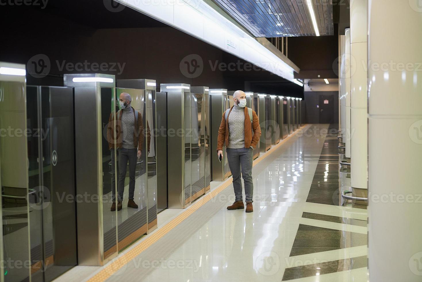 un uomo con una maschera facciale sta usando uno smartphone mentre aspetta un treno della metropolitana foto