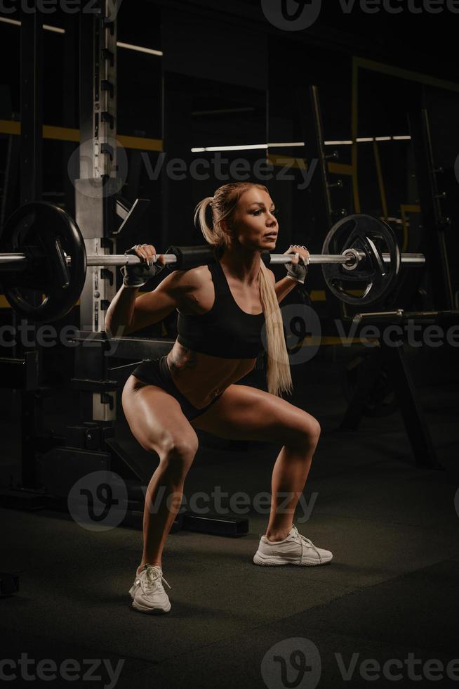 una donna in forma è accovacciata con un bilanciere vicino allo squat rack in una palestra foto