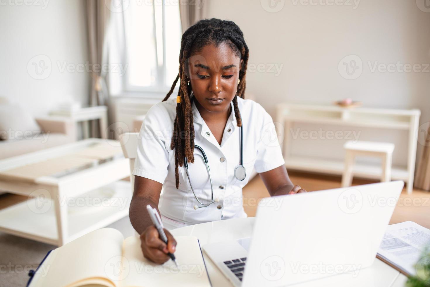 africano professionale femmina medico indossare uniforme assunzione Appunti nel medico rivista, Riempimento documenti, paziente malattia storia, guardare a il computer portatile schermo, alunno Guardando seminario web foto