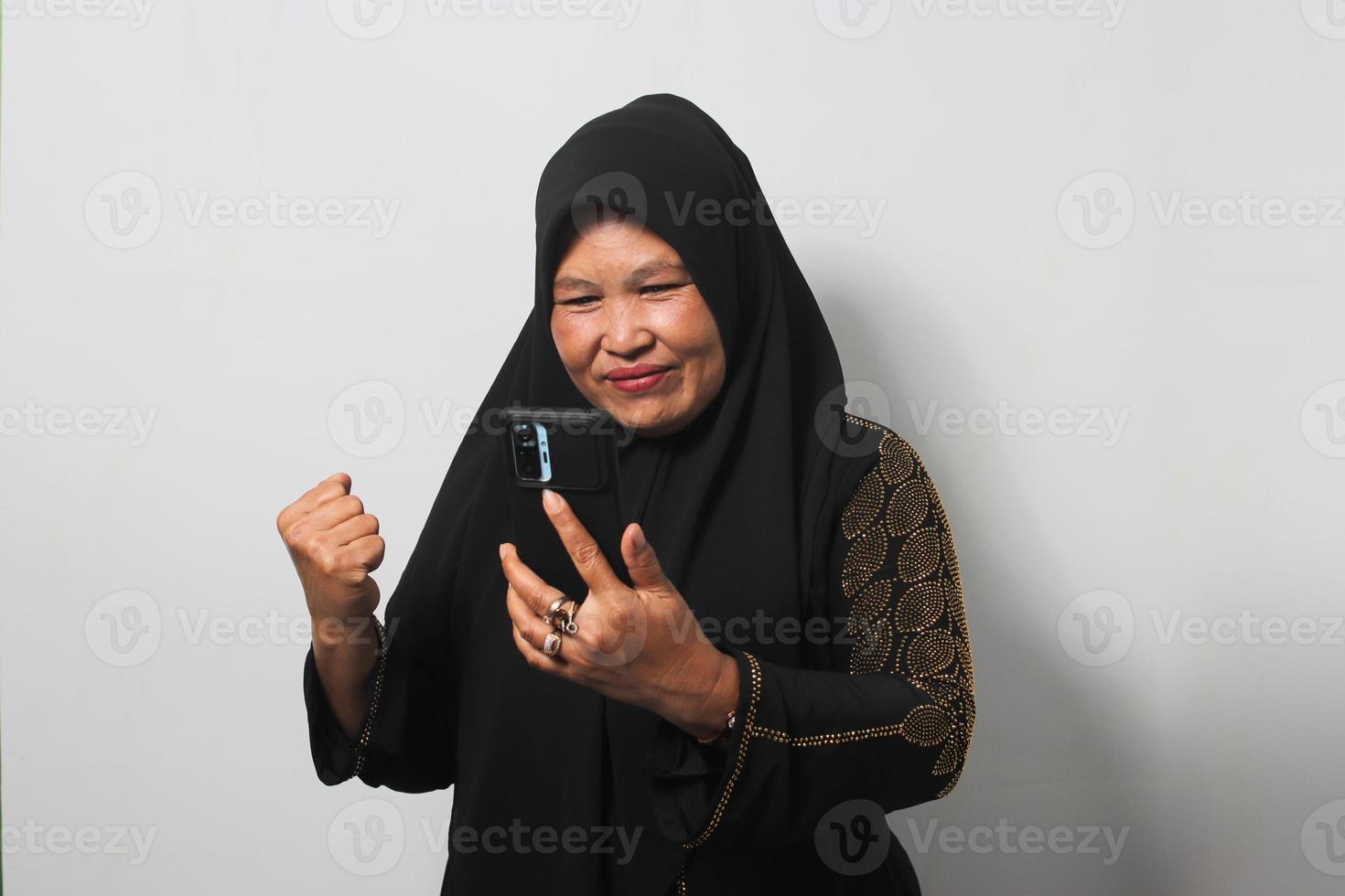 eccitato mezzo anziano asiatico donne indossare hijab guardare a sua Telefono con sì gesto ottenere bene notizia foto