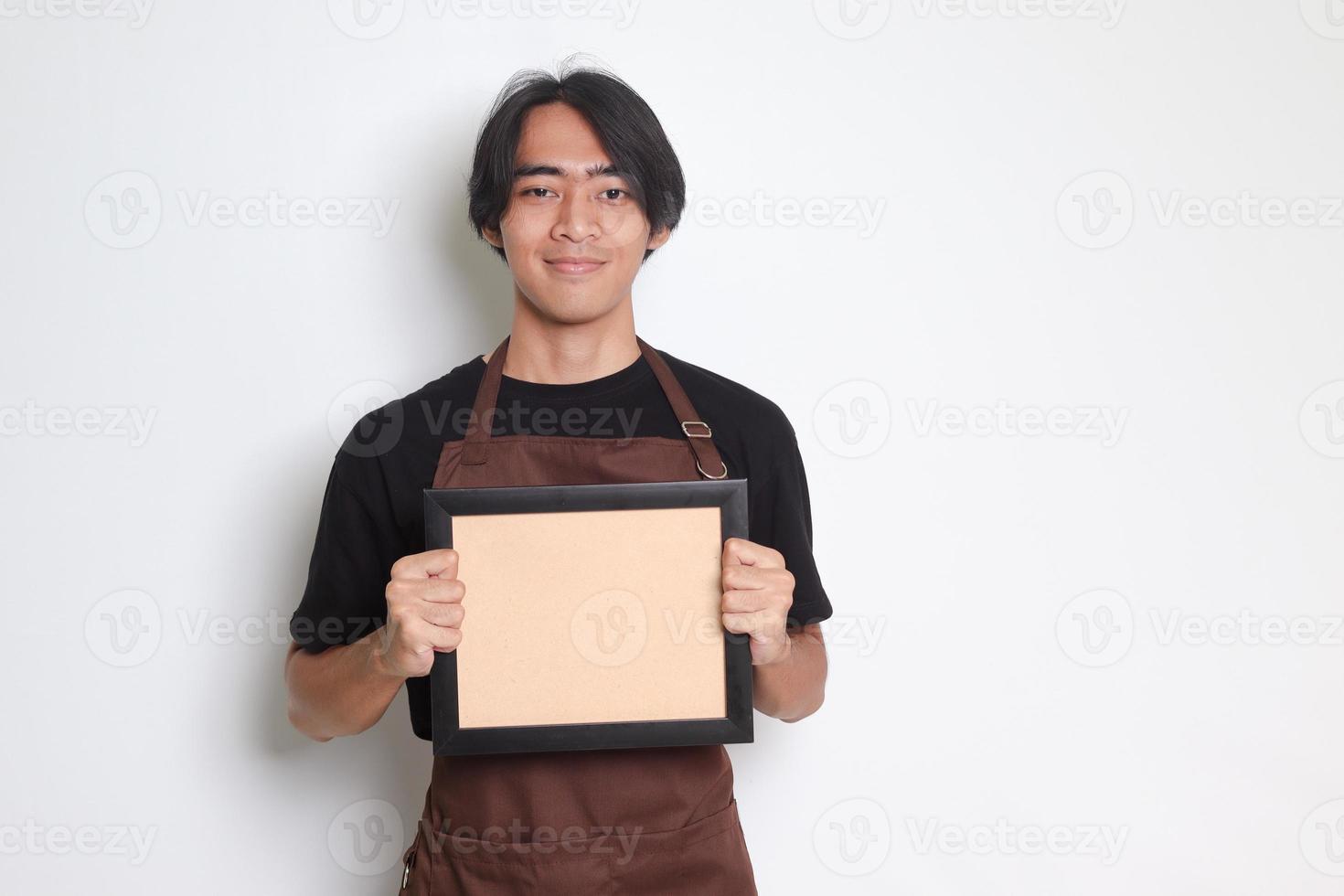 ritratto di attraente asiatico barista uomo nel Marrone grembiule Tenere un vuoto telaio tavola. isolato Immagine su bianca sfondo. foto