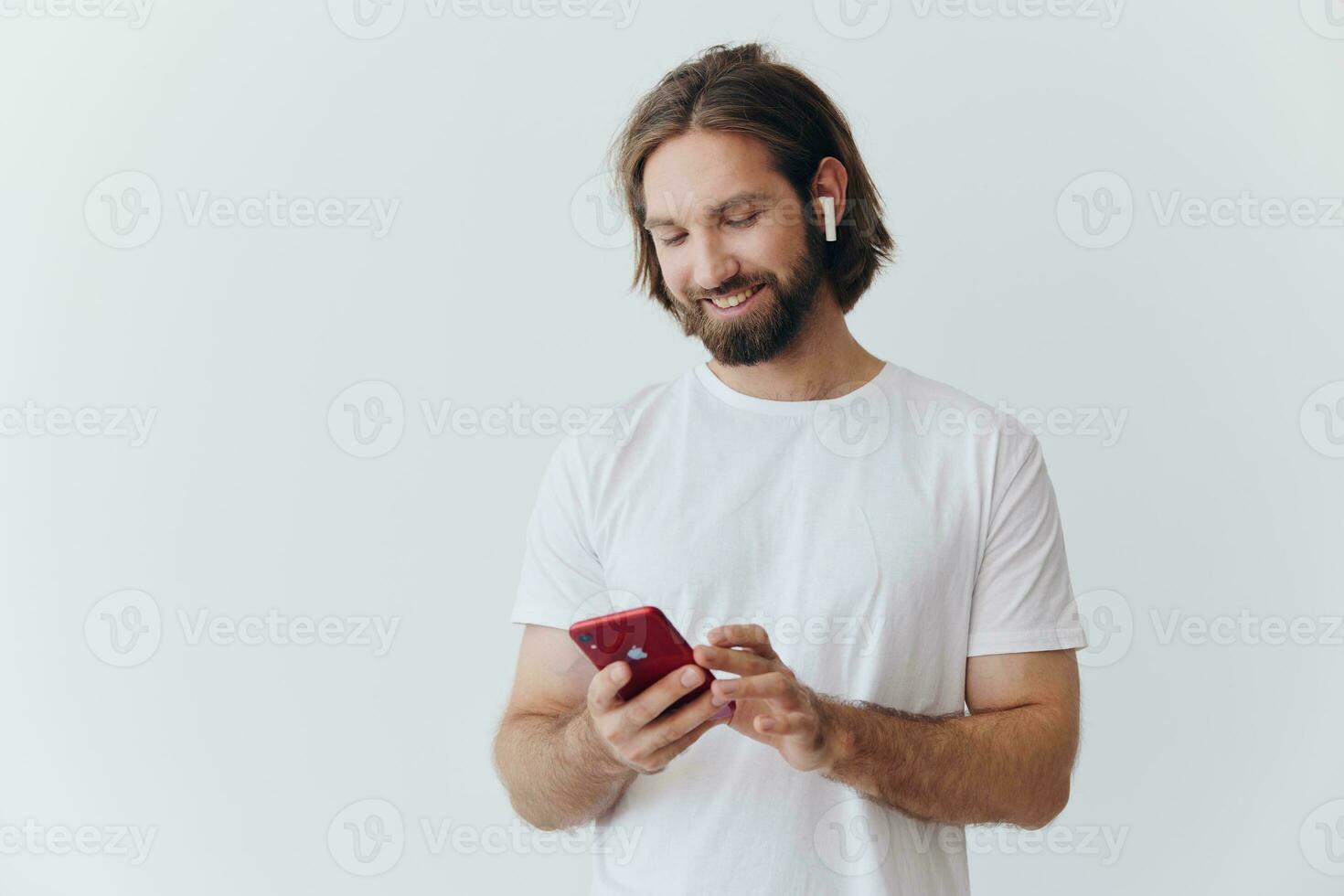 un' uomo con un' barba nel bianca maglietta e jeans sembra a il suo Telefono capovolgimento attraverso un in linea sociale media alimentazione con cuffie nel il suo orecchie ascoltando per un' voce Messaggio su un' bianca sfondo e sorridente foto