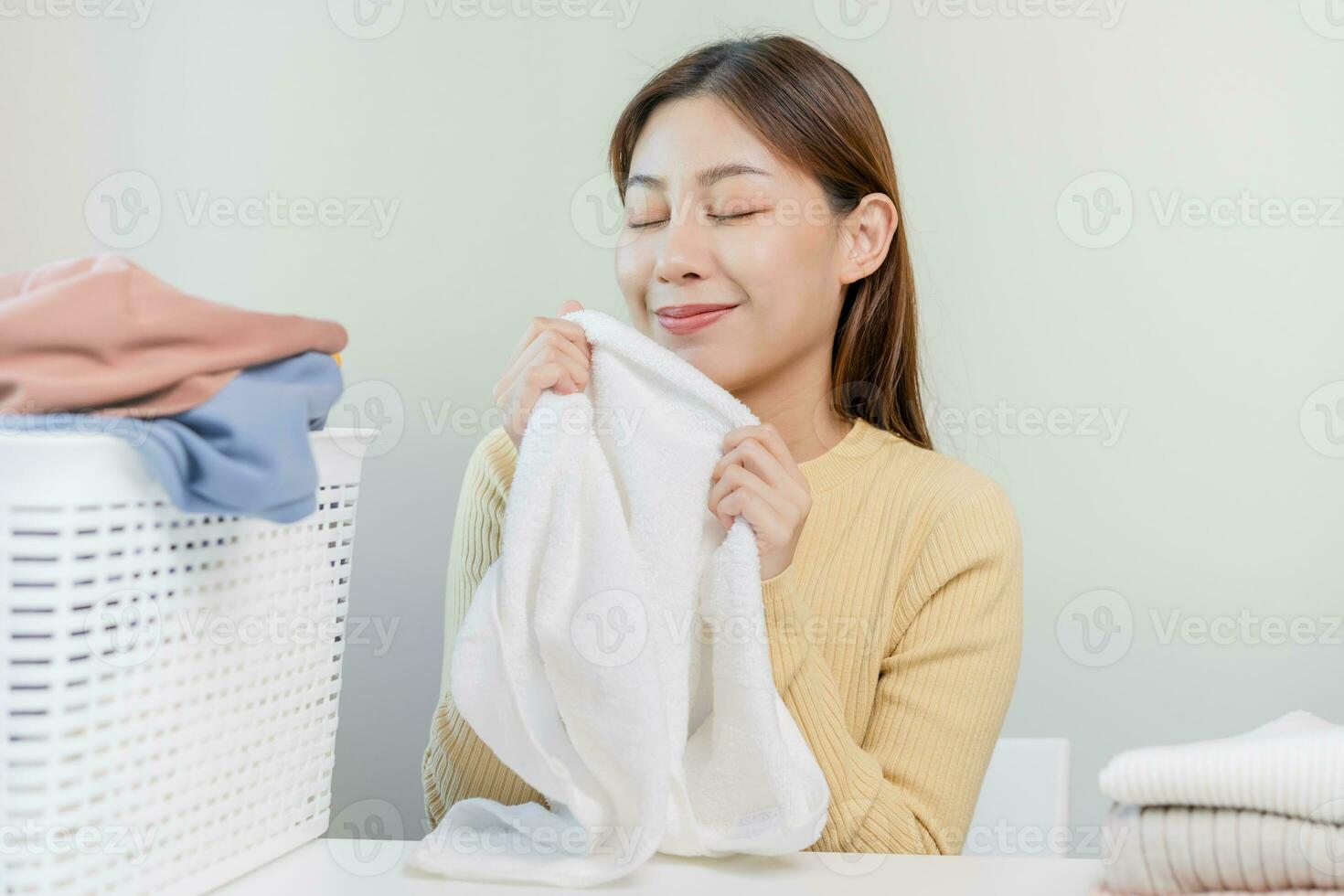 sentire morbidezza, sorridente asiatico giovane donna, ragazza toccante soffice asciugamano cotone, odorare fresco pulito Abiti su tavolo dopo lavaggio, lavanderia, asciutto. domestico Lavorando a casa. lavanderia e domestica. foto