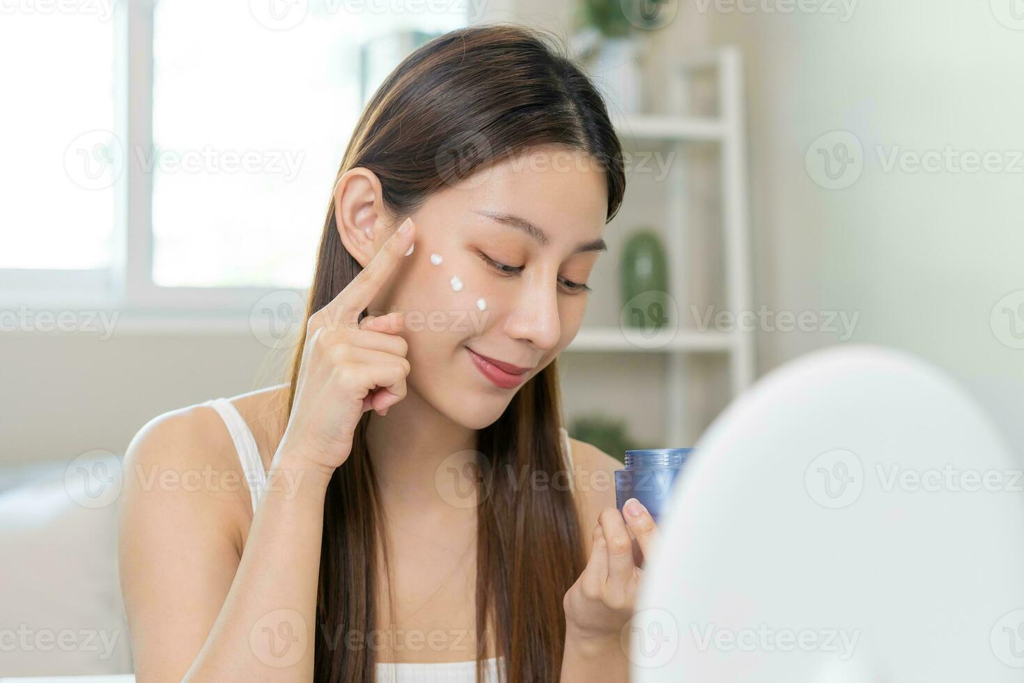 fresco salutare pelle, bellissimo Sorridi di asiatico giovane donna, ragazza guardare a specchio, l'applicazione idratante su sua viso, mettendo crema trattamento prima trucco cosmetico routine a casa. facciale bellezza. foto