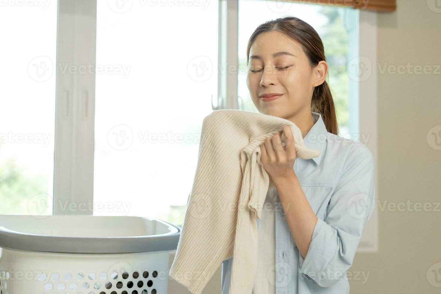 sentire morbidezza, sorridente asiatico giovane donna, ragazza toccante soffice asciugamano cotone, odorare fresco pulito Abiti su tavolo dopo lavaggio, lavanderia, asciutto. domestico Lavorando a casa. lavanderia e domestica. foto