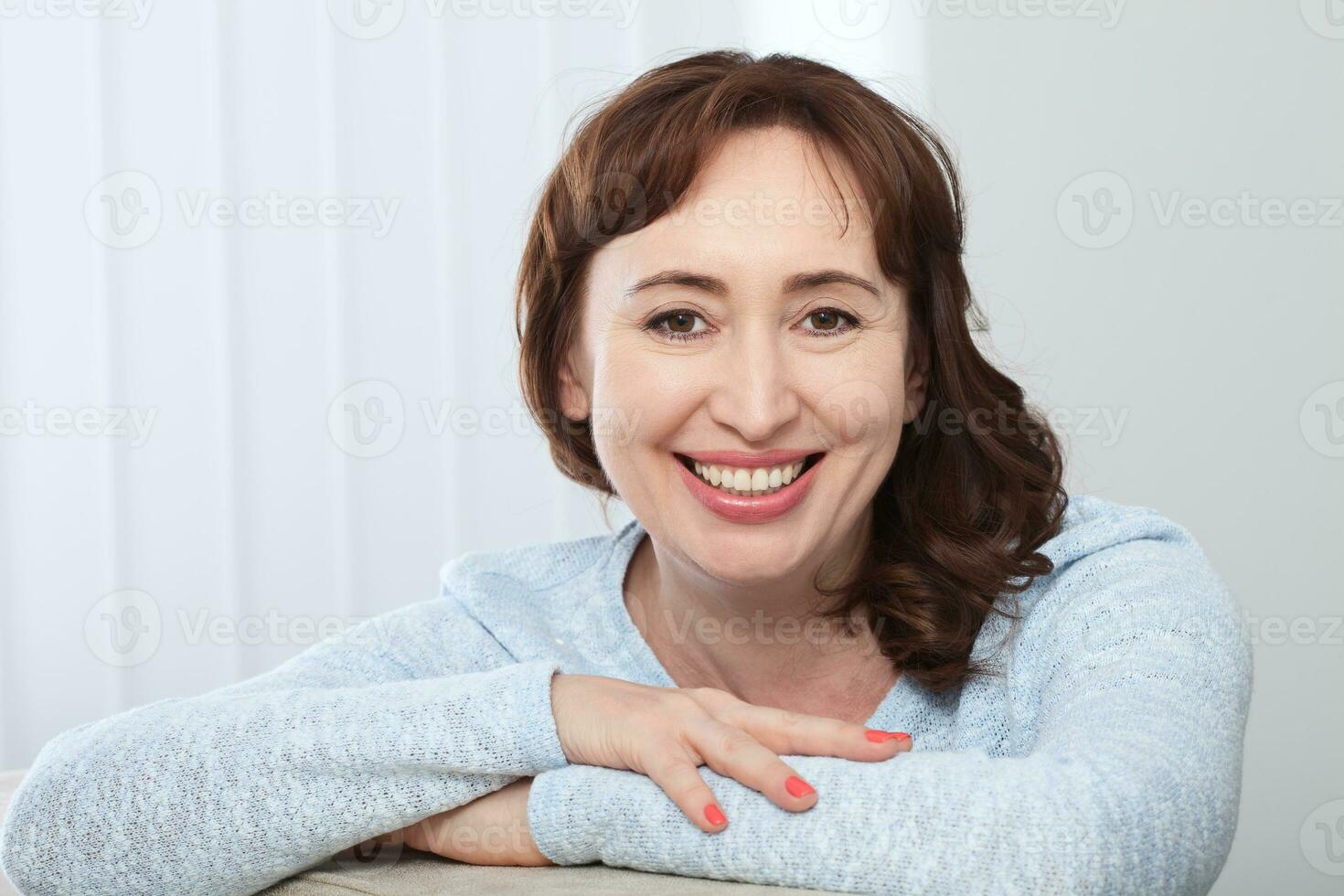 bello di mezza età brunetta donna con un' splendente Sorridi seduta su un' divano a casa guardare a il telecamera foto