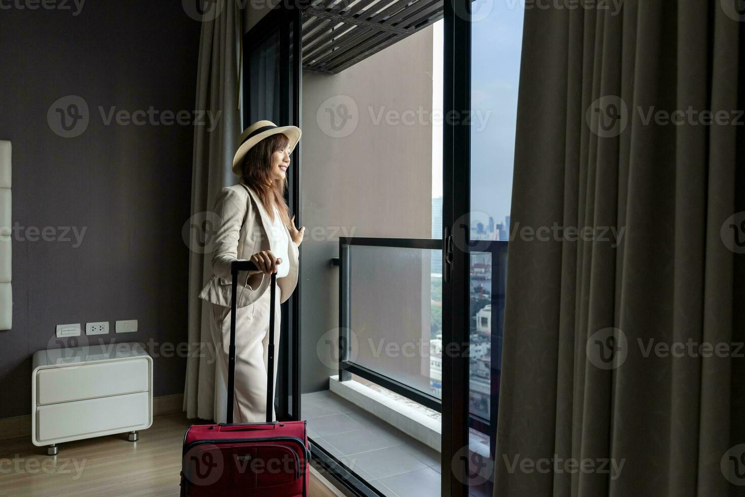 asiatico donna in movimento il lusso Hotel con bagaglio su sua vacanza mentre guardare a il Visualizza a partire dal finestra con alto salire edificio nel il urbano centro la zona per attività commerciale viaggio e condominio vivente uso foto