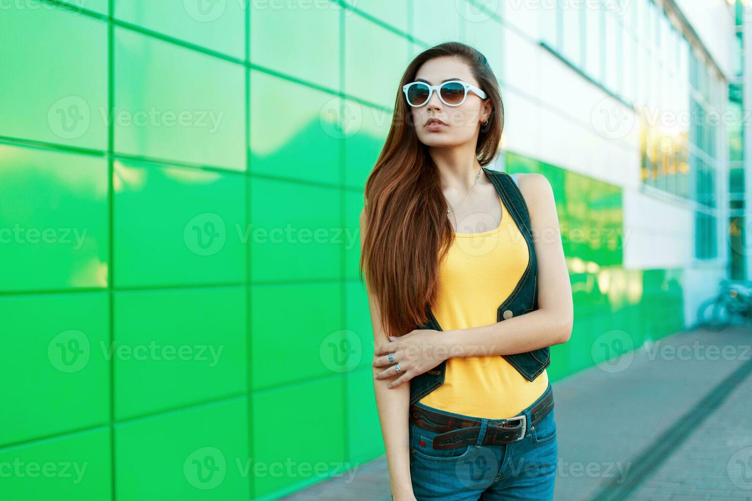 elegante alla moda donna nel occhiali da sole in piedi vicino un' verde costruzione. foto