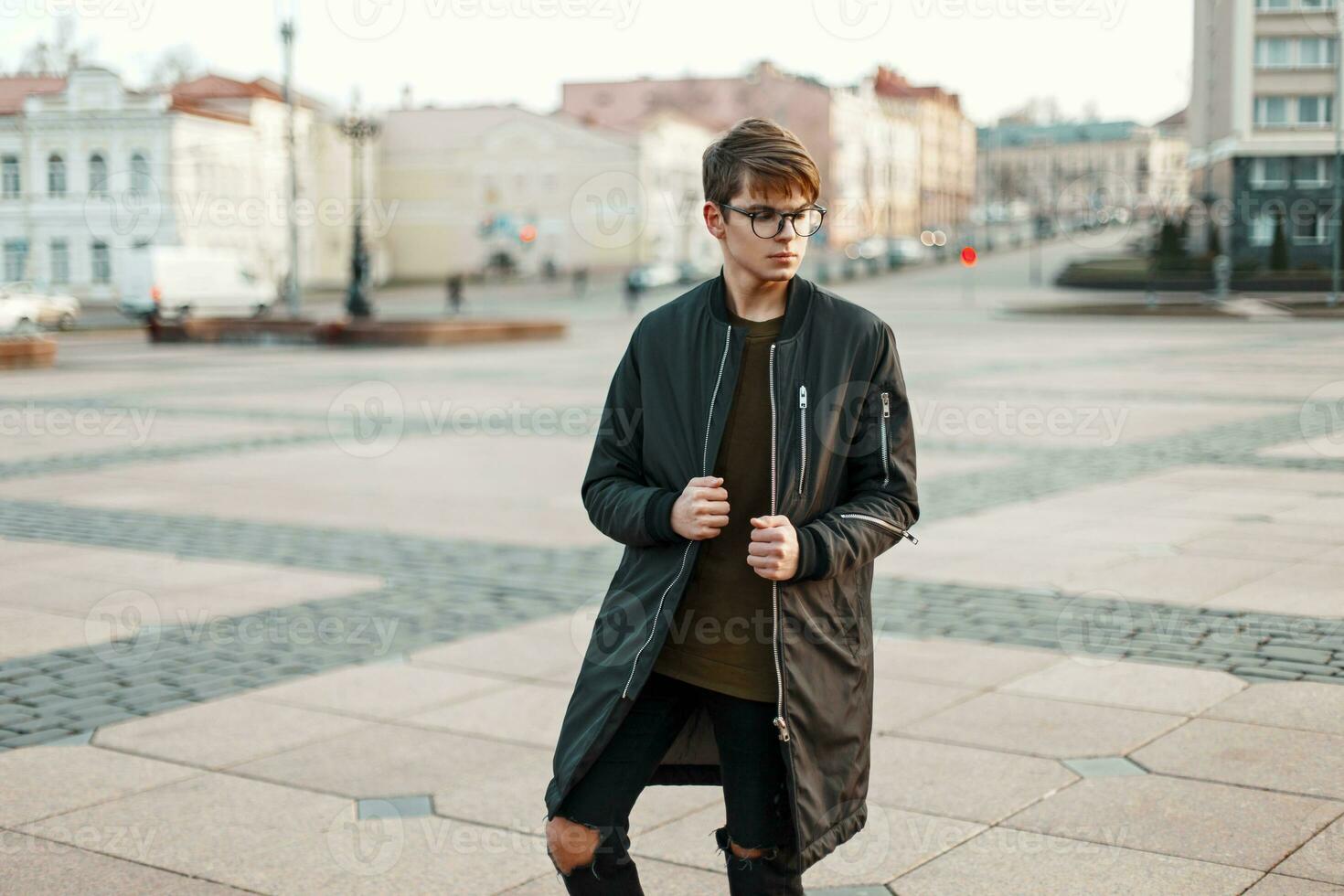 elegante tipo con bicchieri nel di moda giacca e strappato jeans con nero scarpe da ginnastica nel il città foto