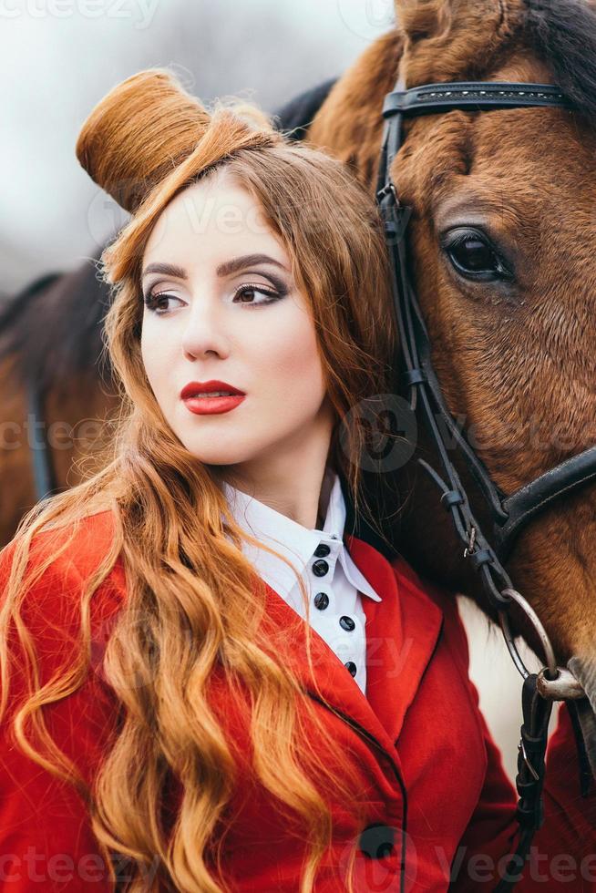 ragazza fantino dai capelli rossi in un cardigan rosso e stivali alti neri con un cavallo foto