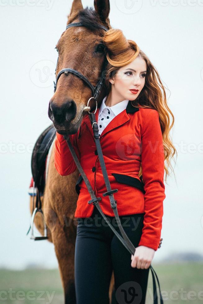 ragazza fantino dai capelli rossi in un cardigan rosso e stivali alti neri con un cavallo foto