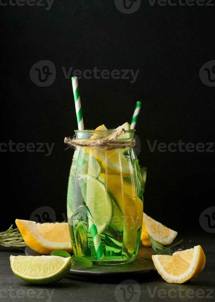 limonata nel un' trasparente bicchiere con Limone, lime, rosmarino rametti e menta le foglie su un' nero sfondo foto