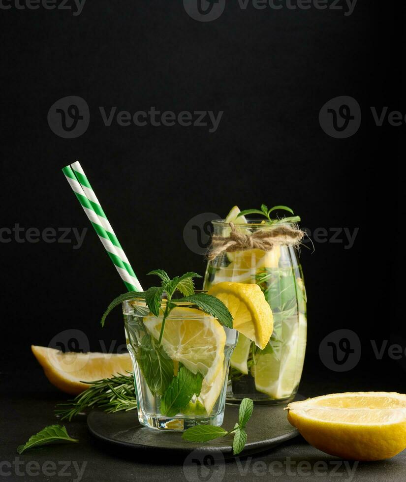 limonata nel un' trasparente bicchiere con Limone, lime, rosmarino rametti e menta le foglie su un' nero sfondo foto