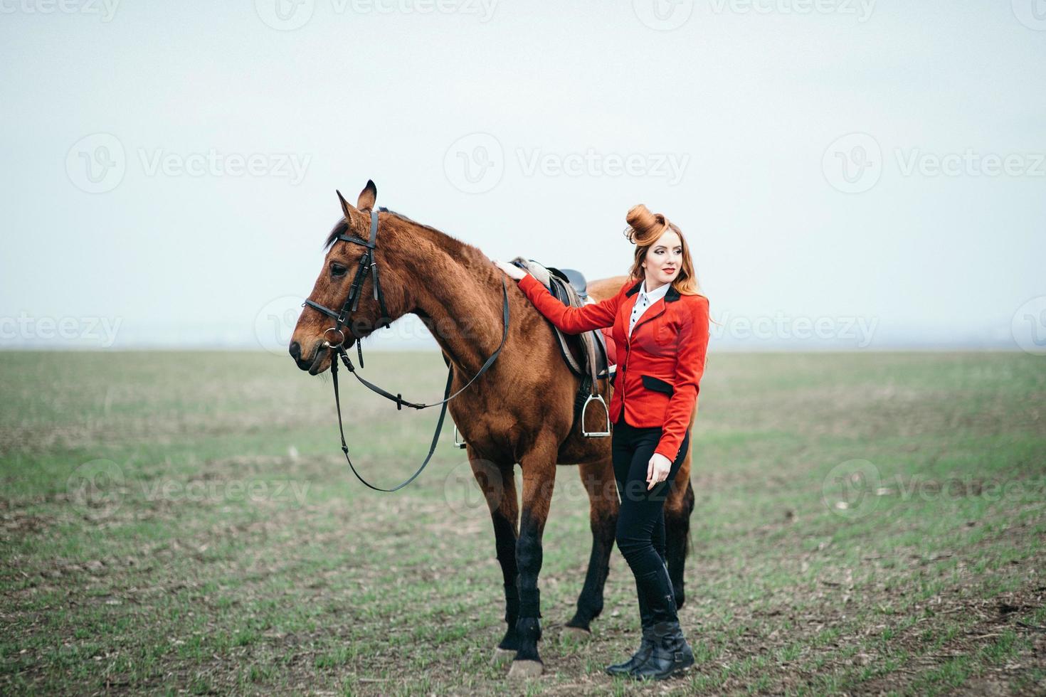 ragazza fantino dai capelli rossi in un cardigan rosso e stivali alti neri con un cavallo foto