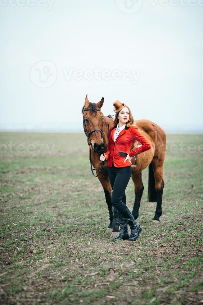 ragazza fantino dai capelli rossi in un cardigan rosso e stivali alti neri con un cavallo foto