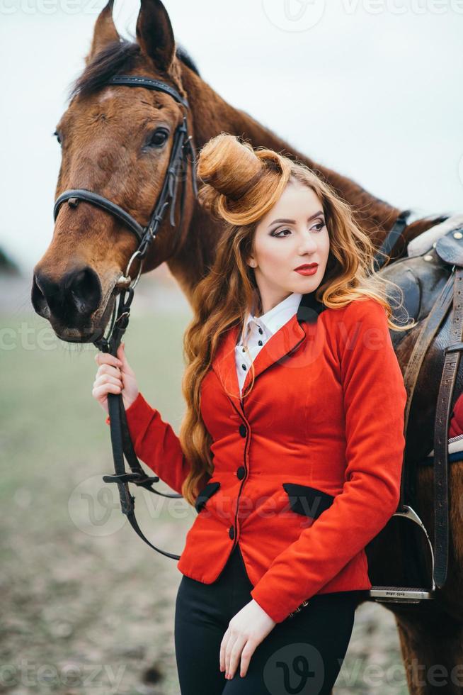 ragazza fantino dai capelli rossi in un cardigan rosso e stivali alti neri con un cavallo foto