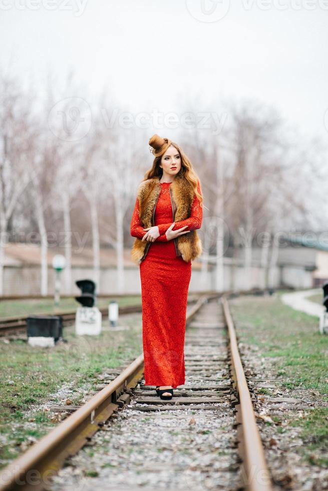 giovane ragazza con i capelli rossi in un abito rosso brillante sui binari ferroviari foto