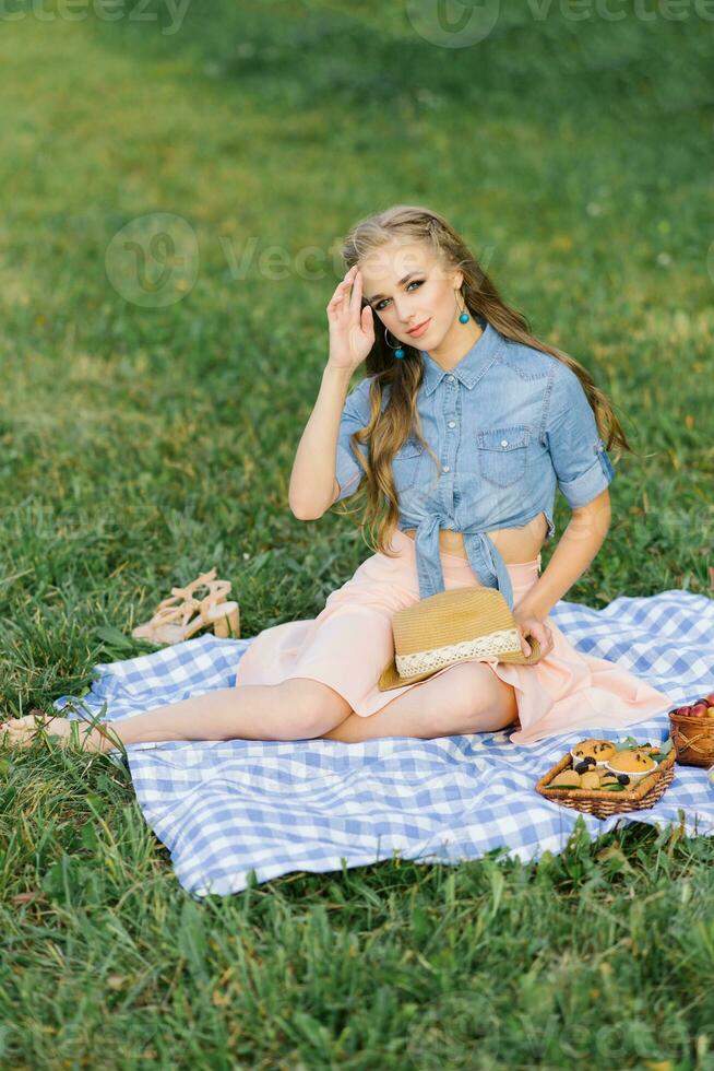 un' bellissimo ragazza sembra contento seduta su un' coperta con frutta quello menzogna su il sfondo di verde erba nel il parco. estate picnic e ricreazione foto