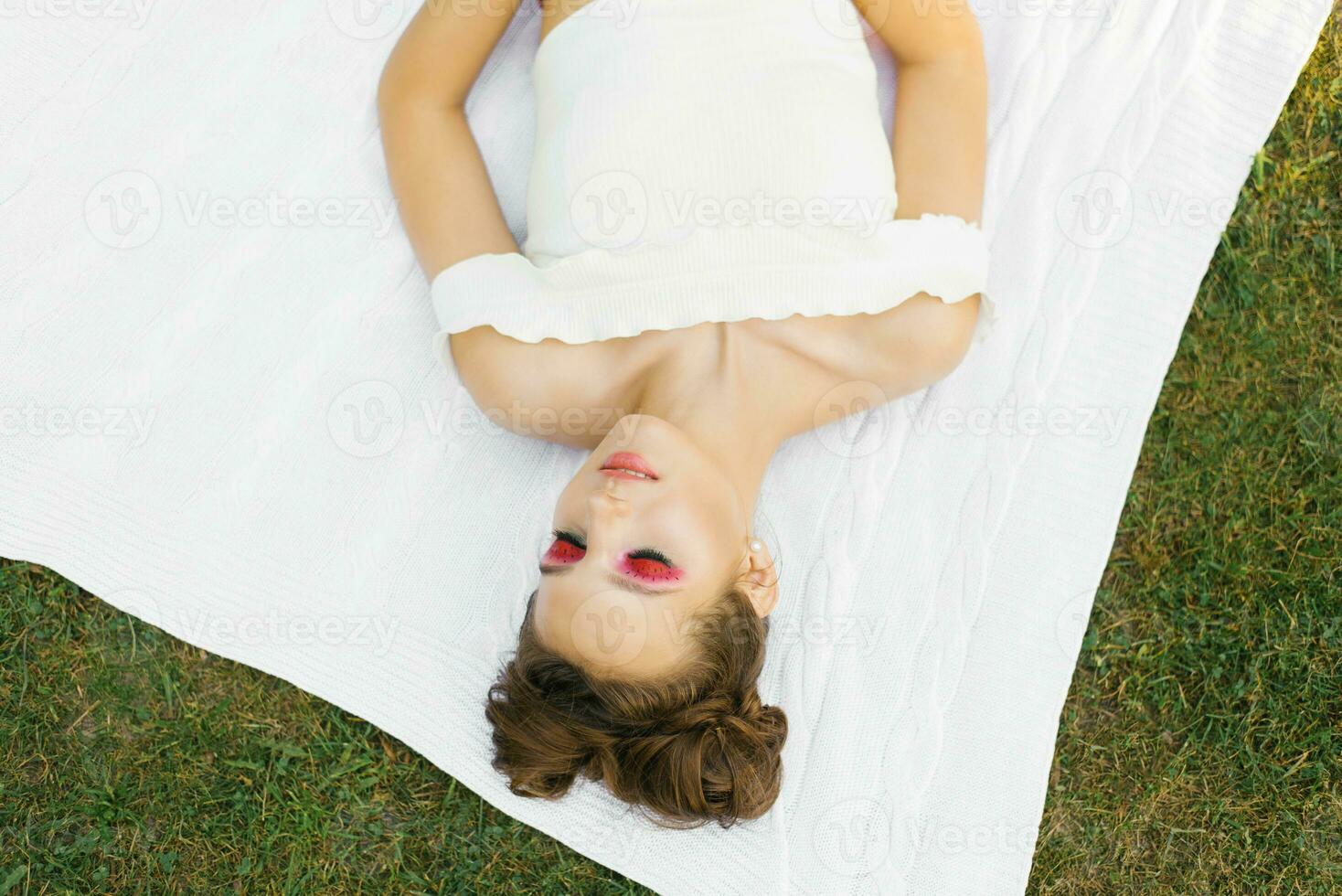 un' bellissimo giovane donna con luminosa trucco nel il stile di anguria bugie con sua occhi chiuso su un' coperta all'aperto nel estate foto