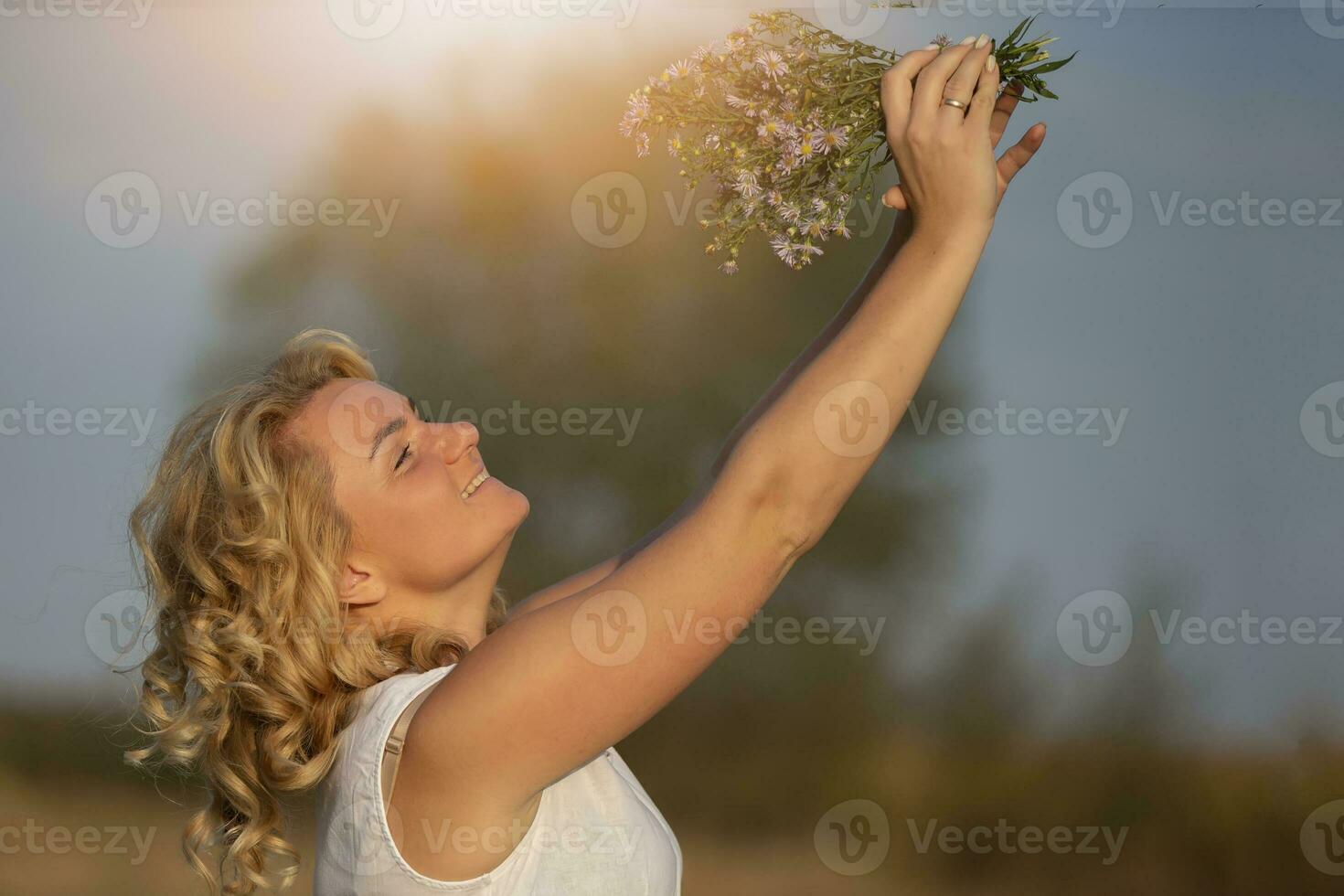 un' bellissimo di mezza età bionda con un' mazzo di fiori selvatici su il sfondo di il sole. foto