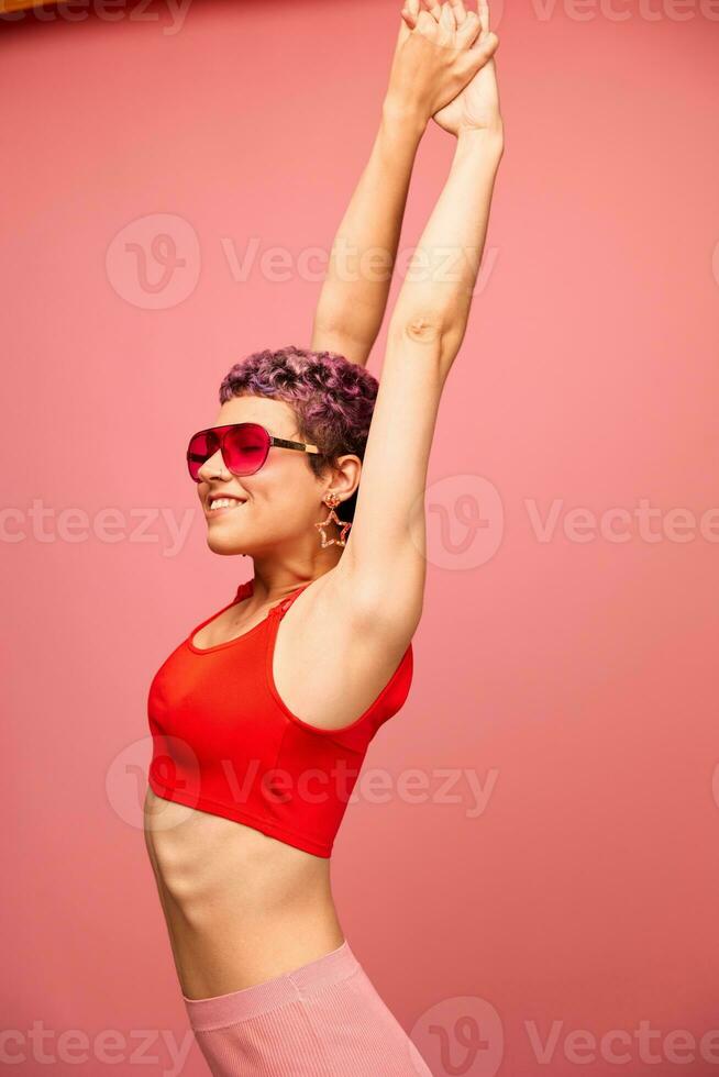 moda ritratto di un' donna con un' corto taglio di capelli nel colorato occhiali da sole con insolito Accessori con orecchini sorridente su un' rosa luminosa sfondo con un' fitness corpo danza foto