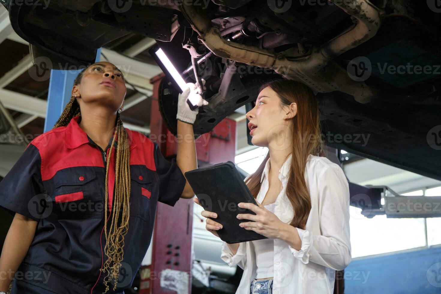 giovane asiatico femmina auto attività commerciale imprenditore discute con meccanico lavoratore, lista di controllo carrello d'atterraggio di sollevato elettrico auto per riparazione a Manutenzione box auto, settore automobilistico servizio lavori industria. foto