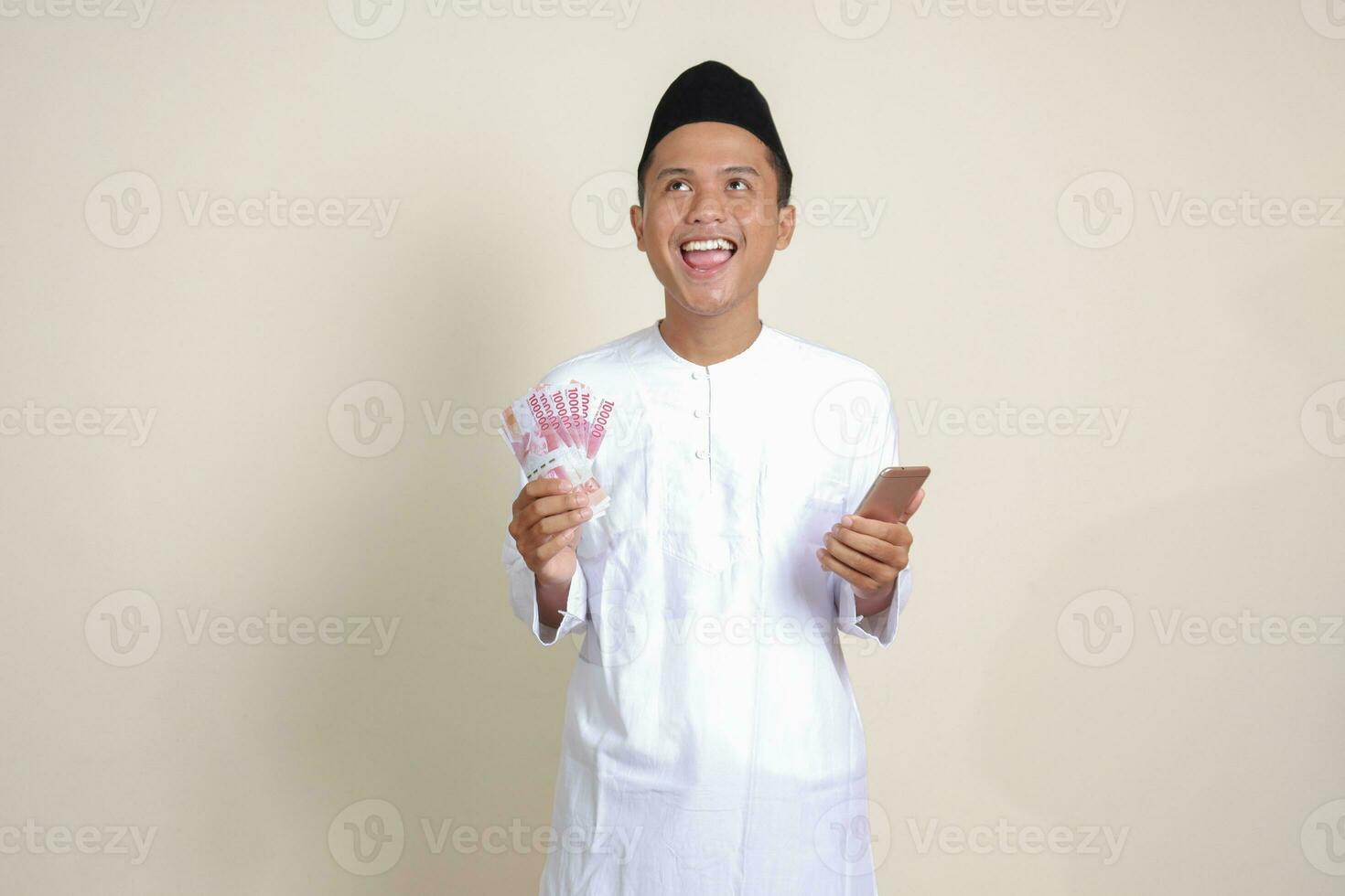 ritratto di attraente asiatico musulmano uomo nel bianca camicia mostrando uno centinaio mille rupia mentre utilizzando mobile Telefono. finanziario e risparmi concetto. isolato Immagine su grigio sfondo foto