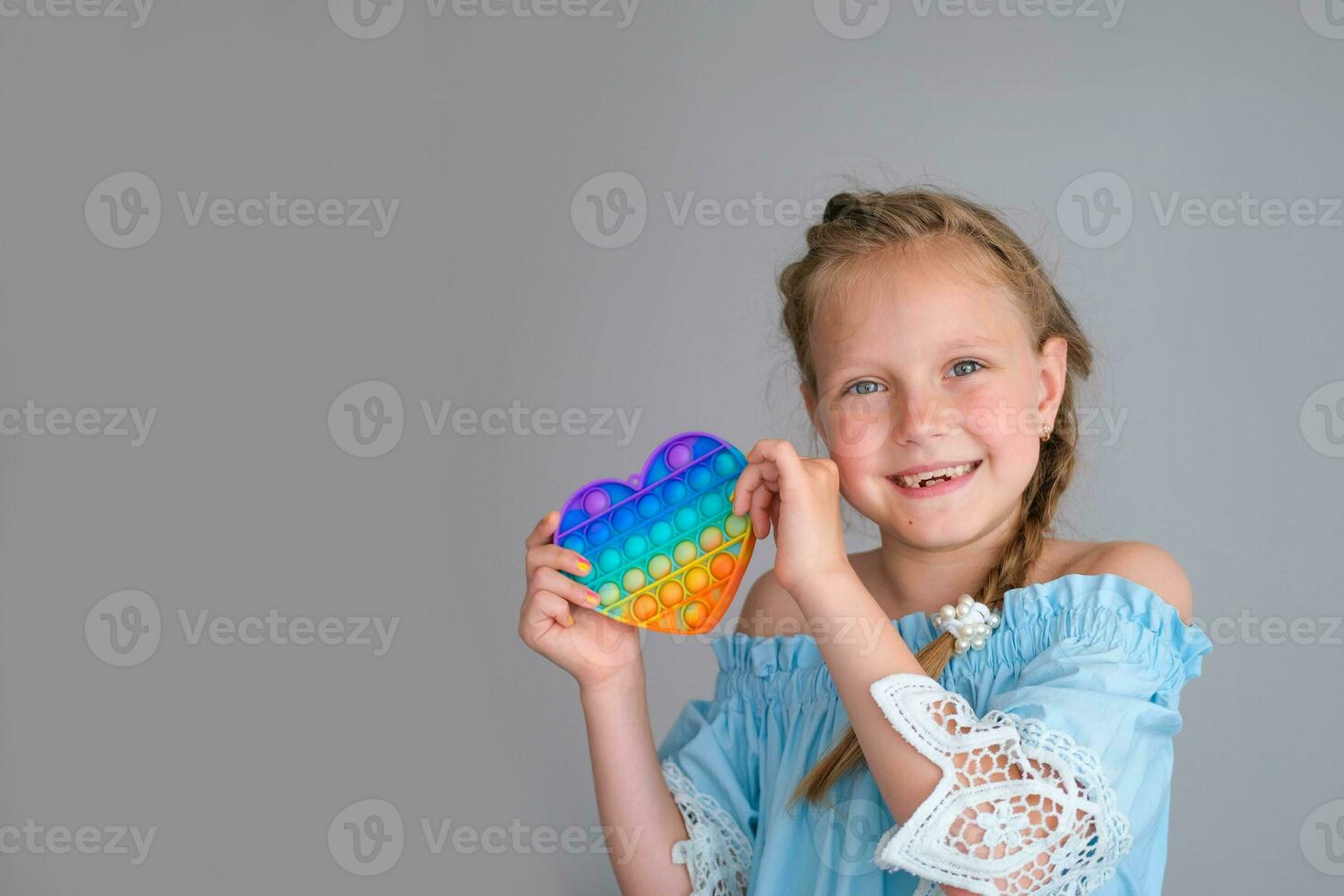 sorridente bambino guardare a il telecamera e Tenere un' arcobaleno pop esso agitarsi giocattolo cuore sagomato. spingere bolla agitarsi sensoriale giocattolo - lavabile e riutilizzabile silicio fatica sollievo giocattolo popit. foto