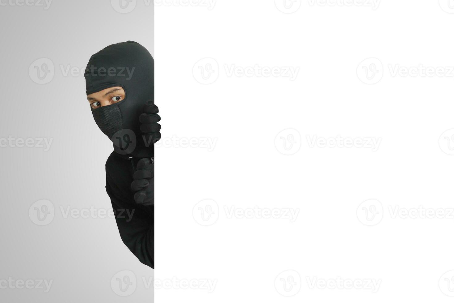 misterioso ladro indossare nero felpa con cappuccio nascondiglio dietro a parete, furtivamente, e guardare per rubato merce. crimine concetto. isolato Immagine su grigio sfondo foto
