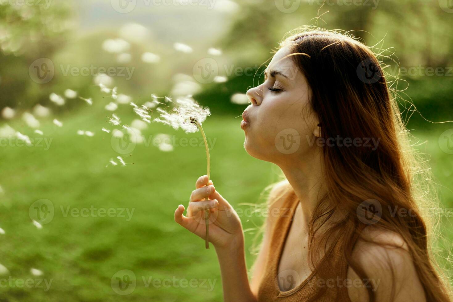 donna con un' dente di leone fiore nel sua mani sorridente e soffiaggio su esso contro un' sfondo di estate verdura e luce del sole foto