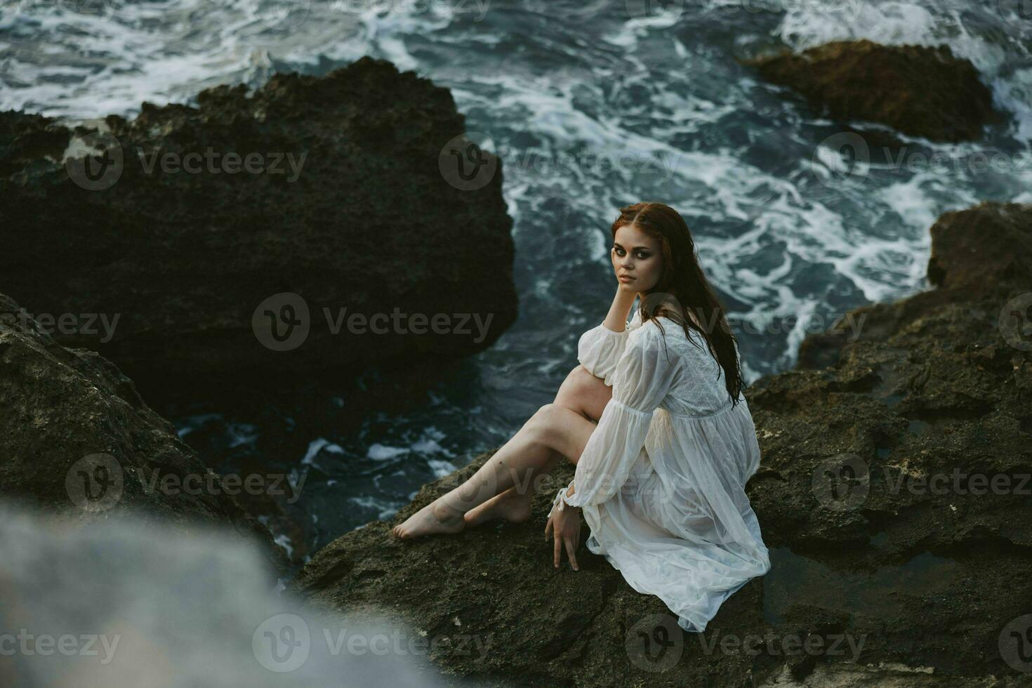 donna con bagnato capelli a piedi nudi nel un' bianca vestito si siede su un' scogliera natura inalterato foto