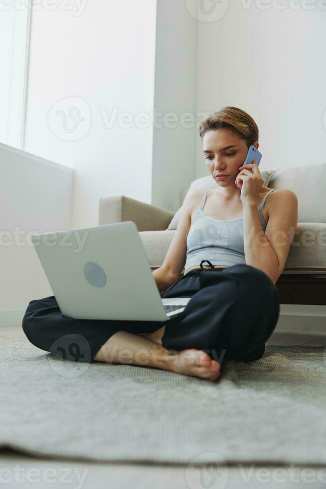 libero professionista donna con il computer portatile e Telefono lavori a partire dal casa seduta su il pavimento nel sua casa Abiti con un' corto taglio di capelli, gratuito copia spazio foto