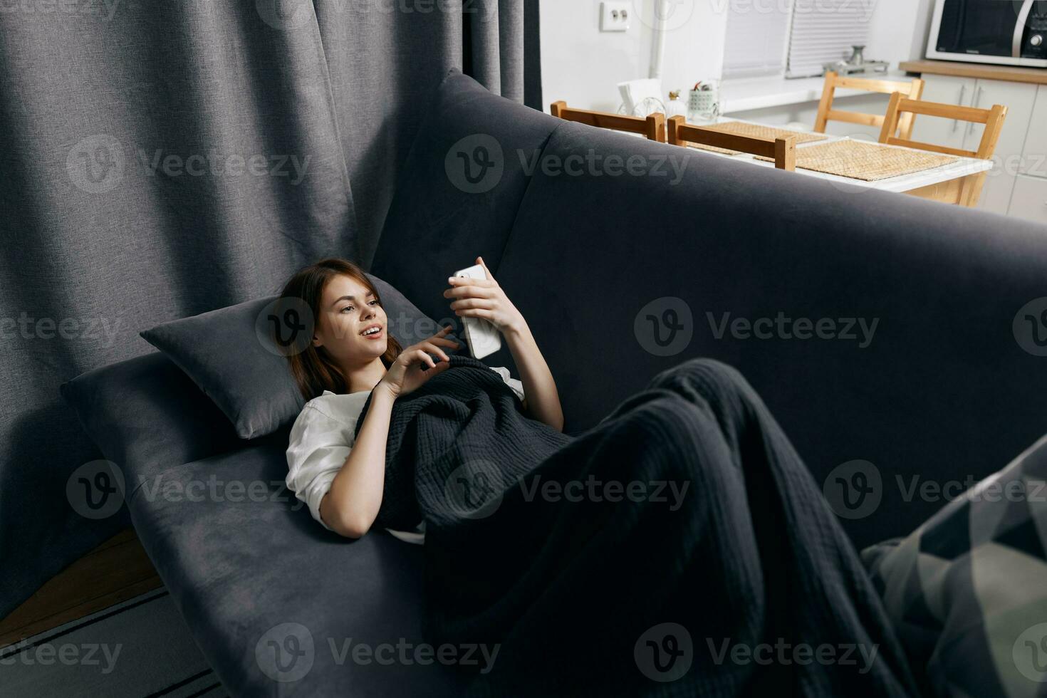 giovane donna coperto con un' coperta bugie su il divano con un' mobile Telefono nel sua mano foto